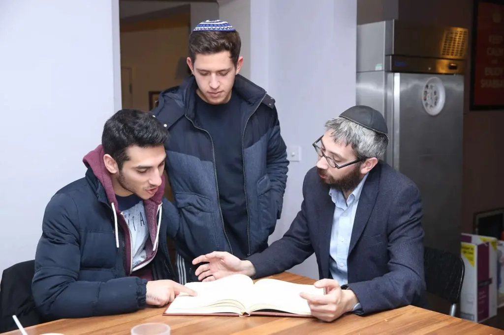 A rabbi and two college students studying a Jewish text together at a table